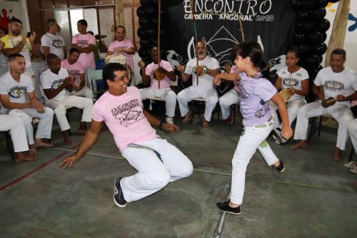 Mestre André dodging while his young student throws a kick. They are playing in front of a group of capoeiristas, some of them playing musical instruments.