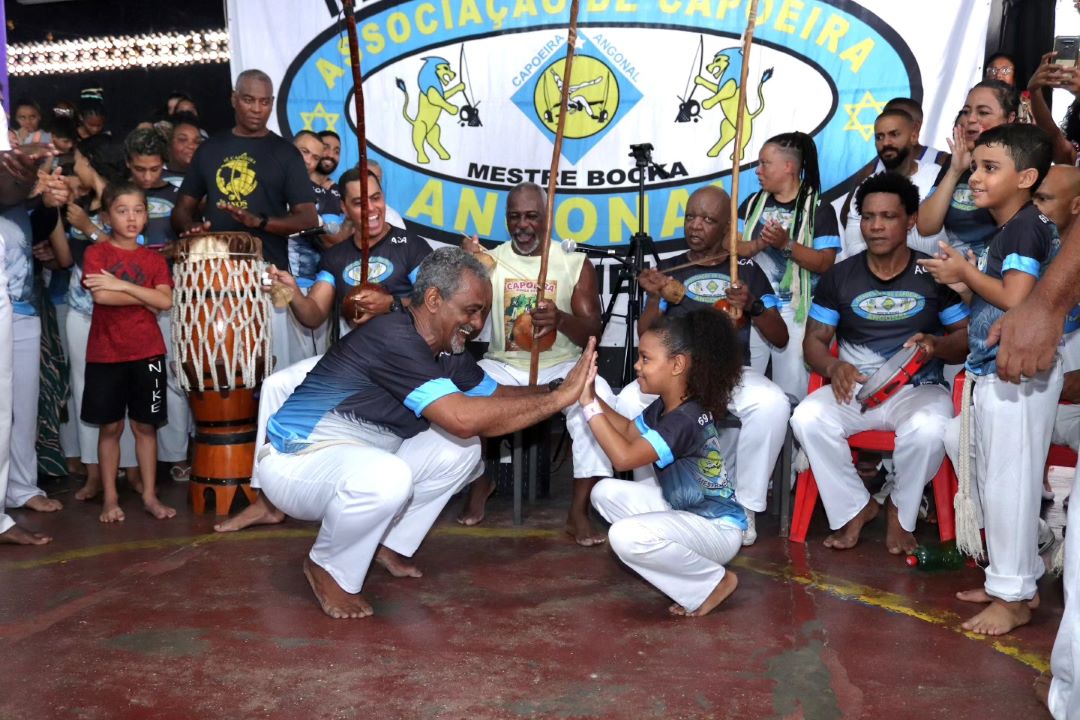 Mestre Bocka and his grand-daughter crouch facing each other, giving one another a 'high ten' and smiling as they prepare to play Capoeira. Behind them is a group of people playing Capoeira musical instruments.